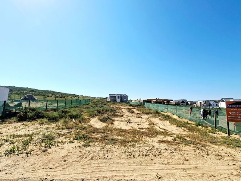 A vacant residential plot featuring sandy terrain and perimeter fencing, situated in a developing coastal neighborhood.