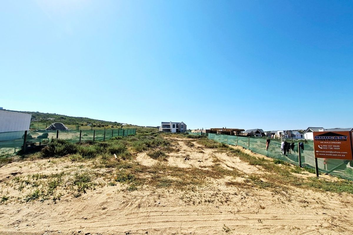 A vacant residential plot featuring sandy terrain and perimeter fencing, situated in a developing coastal neighborhood.