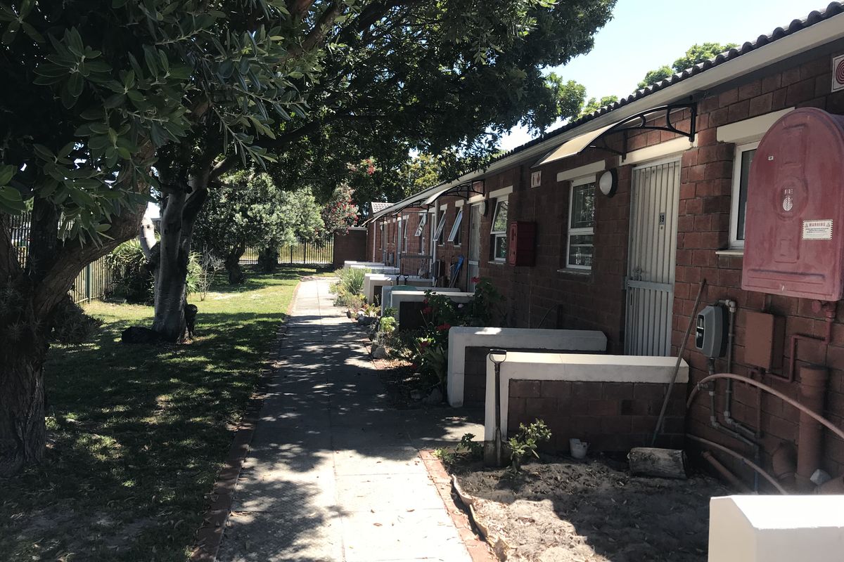 A view of the rear exterior of the townhouse complex, featuring a paved walkway, brick facade, and established trees along the garden area.
