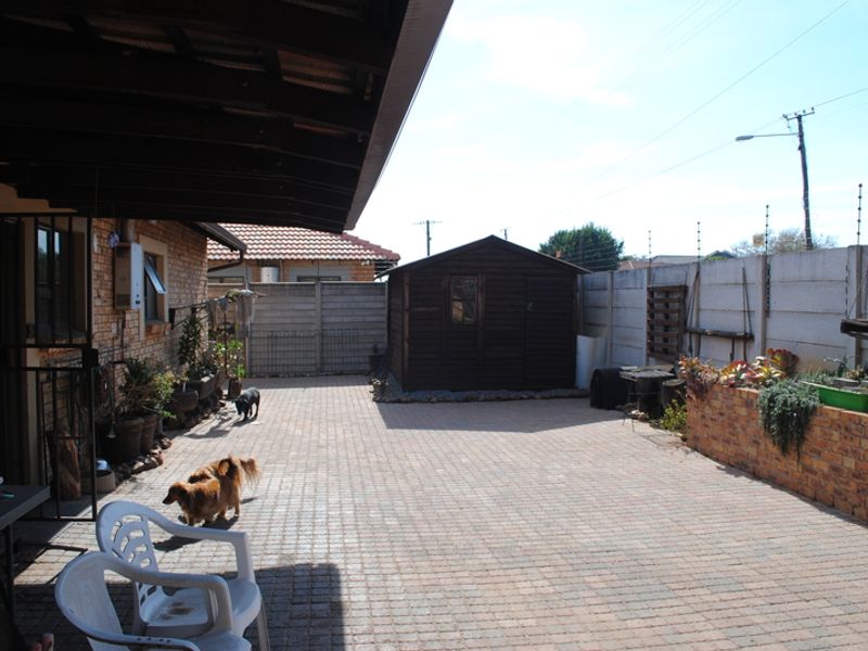 A paved rear courtyard featuring a covered patio area, a timber garden shed, and a brick retaining wall enclosed by a secure boundary wall.