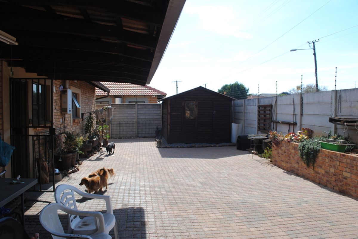 A paved rear courtyard featuring a covered patio area, a timber garden shed, and a brick retaining wall enclosed by a secure boundary wall.