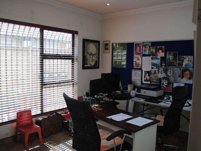 A functional home office space featuring tiled flooring, recessed downlighting, and large windows fitted with security bars and venetian blinds.