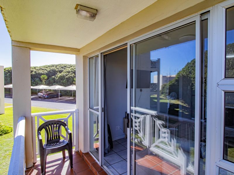 A covered balcony featuring tiled flooring, a sliding glass door entrance, and a protective railing, overlooking the complex parking area.