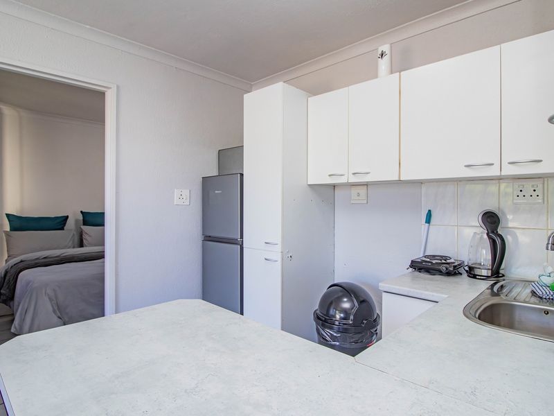 Compact kitchen featuring white built-in cabinetry, laminate countertops, and a stainless steel sink, with an adjacent view into the bedroom.