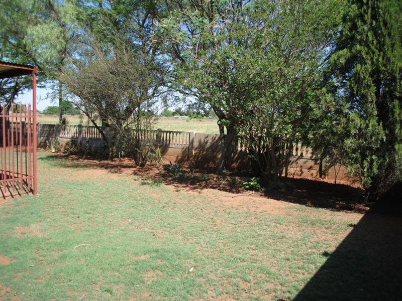 A private backyard area featuring a lawn, mature trees, and a precast concrete boundary wall with metal security fencing.