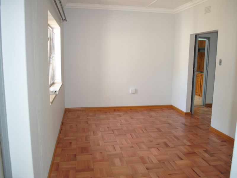 A bright, empty bedroom featuring well-maintained parquet flooring, neutral walls, and security bars on the window.