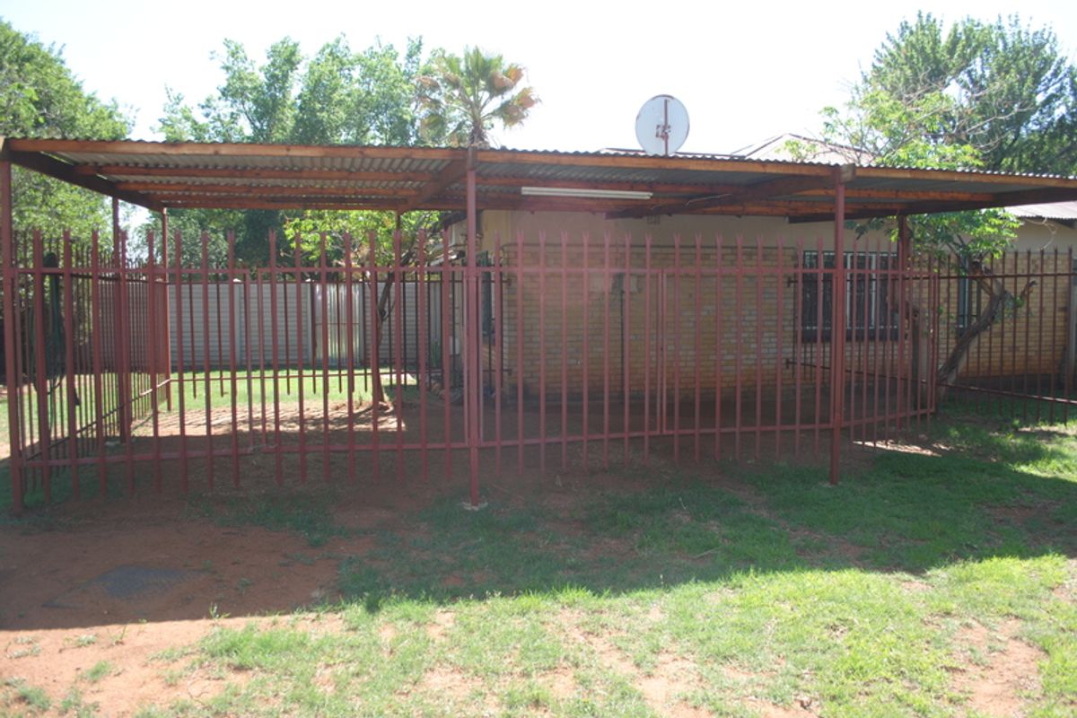 Rear exterior view showing a covered patio area with a corrugated metal roof, steel support structure, and perimeter security fencing.
