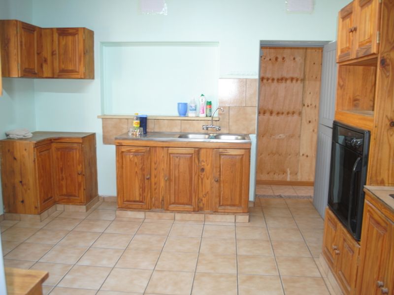 A dated kitchen featuring wooden cabinetry, tiled flooring, and a double basin sink, with an adjacent room showing unfinished wall surfaces.