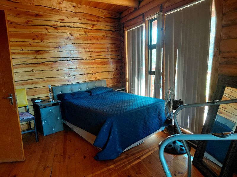 A rustic bedroom featuring natural log walls, timber flooring, and exposed ceiling beams with large windows fitted with vertical blinds.