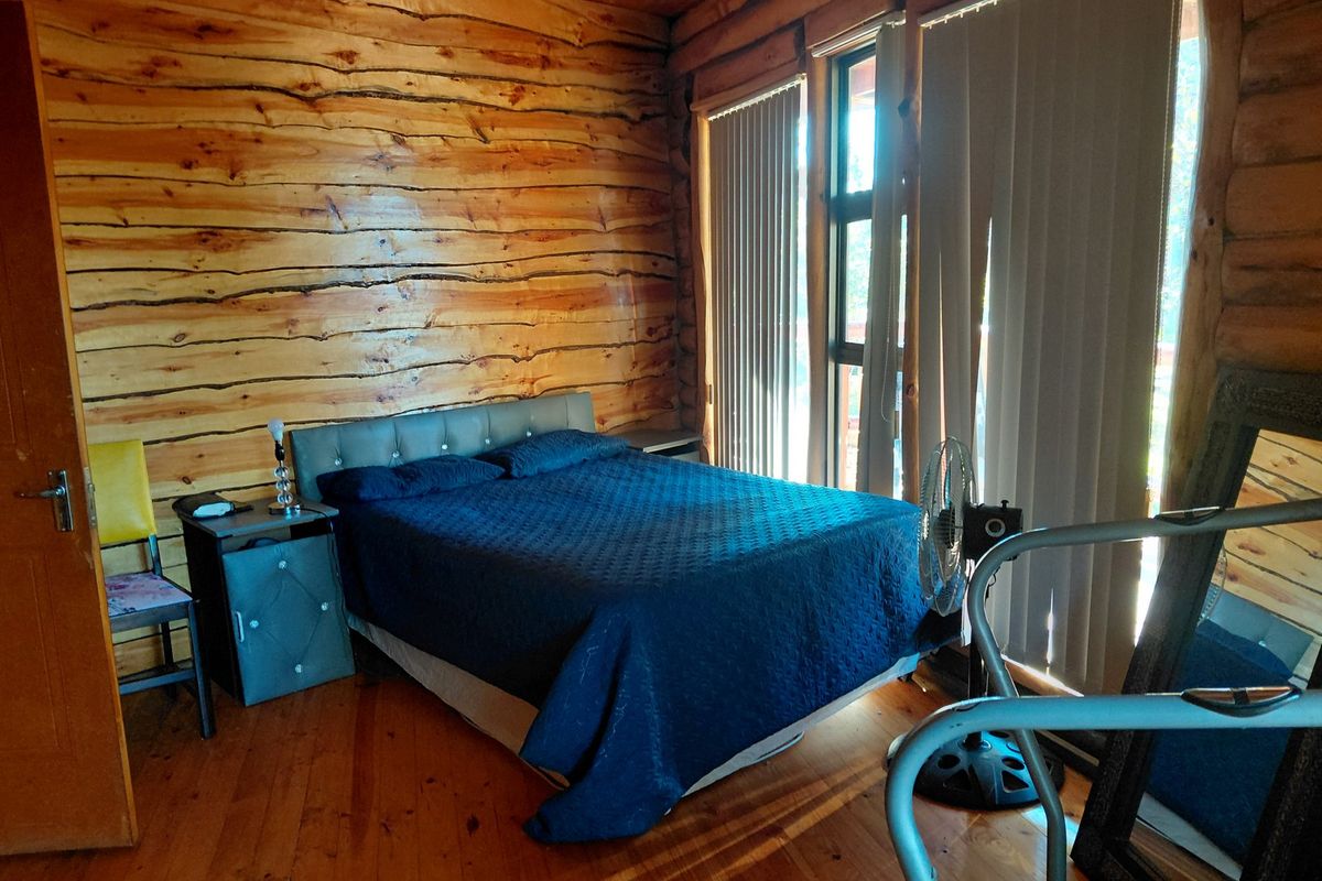 A rustic bedroom featuring natural log walls, timber flooring, and exposed ceiling beams with large windows fitted with vertical blinds.