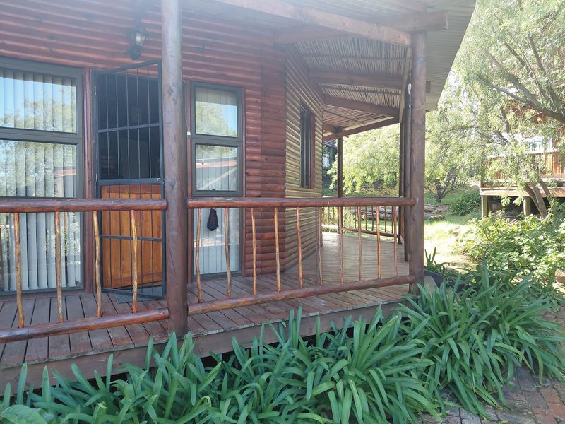 A rustic timber log cabin featuring a wrap-around wooden deck, security gate, and thatched roof overhang, set within a natural garden environment.