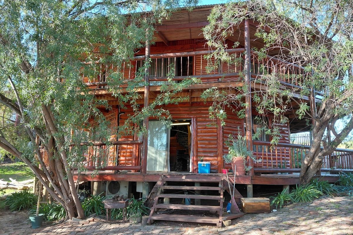 A two-story timber log cabin featuring a wrap-around balcony, wooden decking, and stairs, set within a natural garden environment.