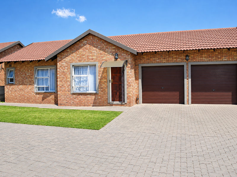 A well-maintained townhouse featuring a face brick facade, double garage, paved driveway, and a neat front lawn under a clear blue sky.
