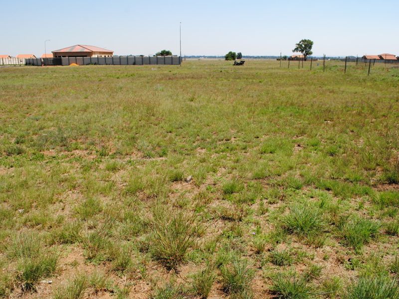A wide, level vacant residential plot featuring natural grass cover and perimeter fencing, situated in a developing residential area.