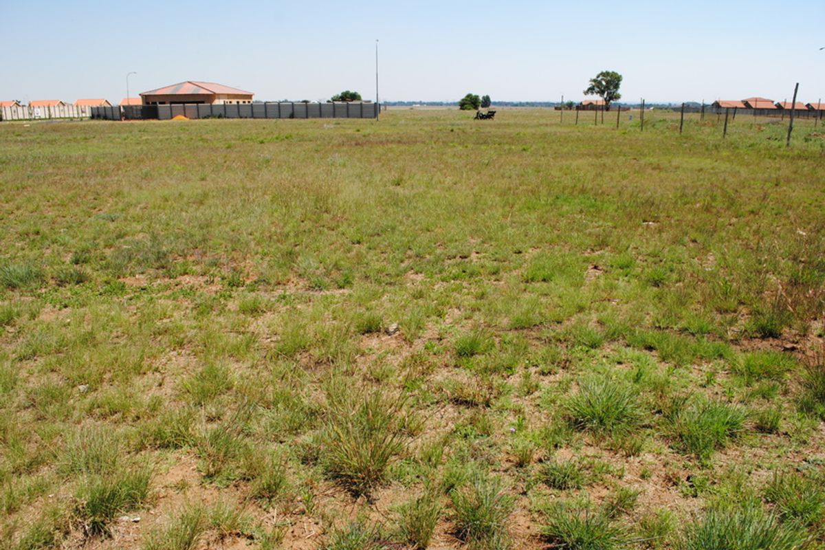 A wide, level vacant residential plot featuring natural grass cover and perimeter fencing, situated in a developing residential area.
