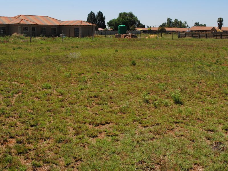 A vacant residential plot featuring natural grass coverage, perimeter wire fencing, and a visible water storage tank in a suburban setting.