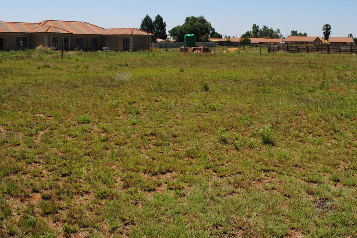 A vacant residential plot featuring natural grass coverage, perimeter wire fencing, and a visible water storage tank in a suburban setting.
