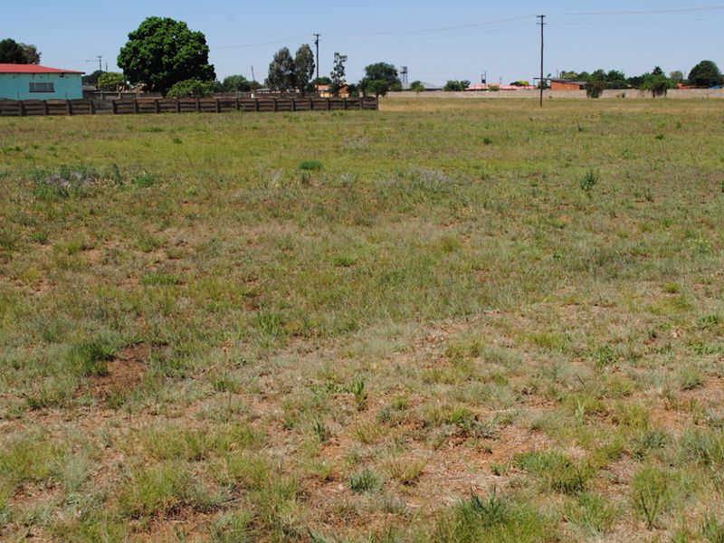 A vacant residential plot of land featuring open grass, perimeter fencing, and nearby utility infrastructure in a suburban setting.