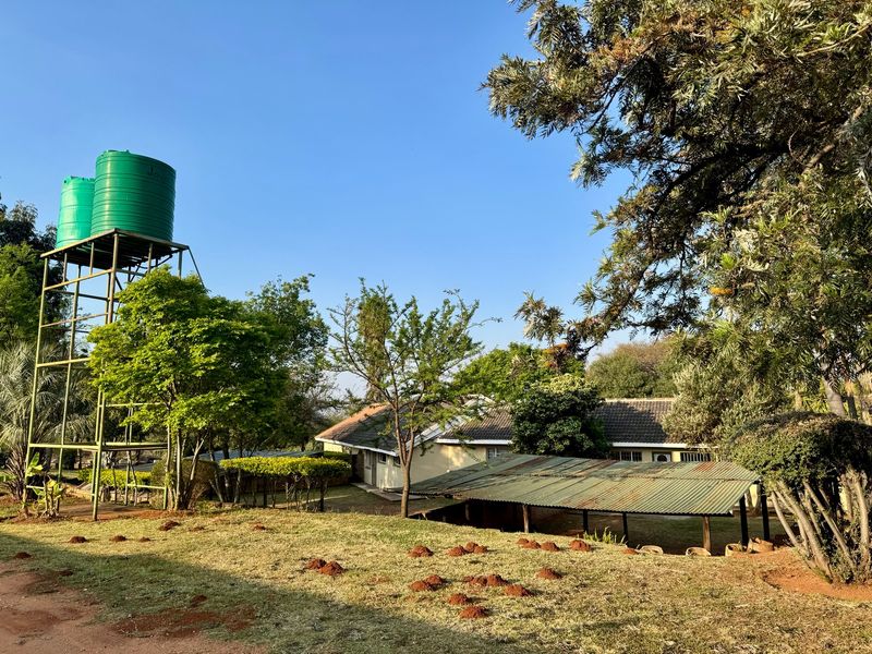 Rear view of the farm property featuring a metal water tank stand, a corrugated roof structure, and established garden surroundings.