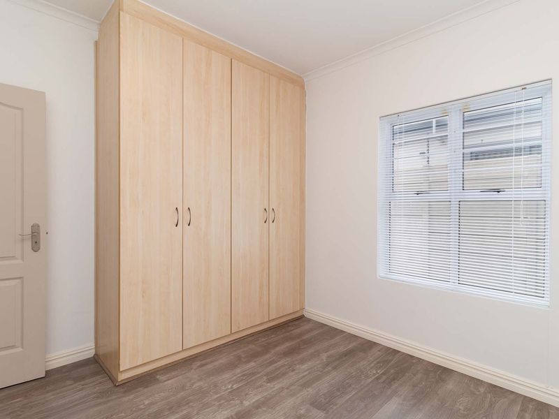 A clean, empty bedroom featuring light wood-finish built-in wardrobes, modern laminate flooring, and a window fitted with venetian blinds.