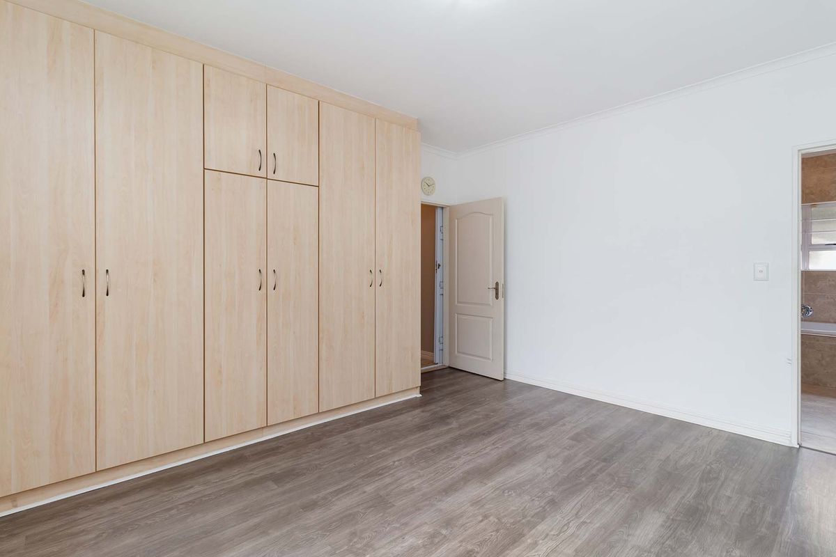 A clean, empty bedroom featuring light wood-finish built-in cupboards, modern laminate flooring, and access to an adjoining bathroom.