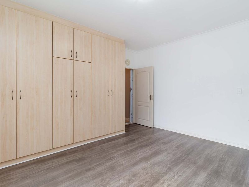 A clean, empty bedroom featuring light wood-finish built-in cupboards, modern laminate flooring, and access to an adjoining bathroom.