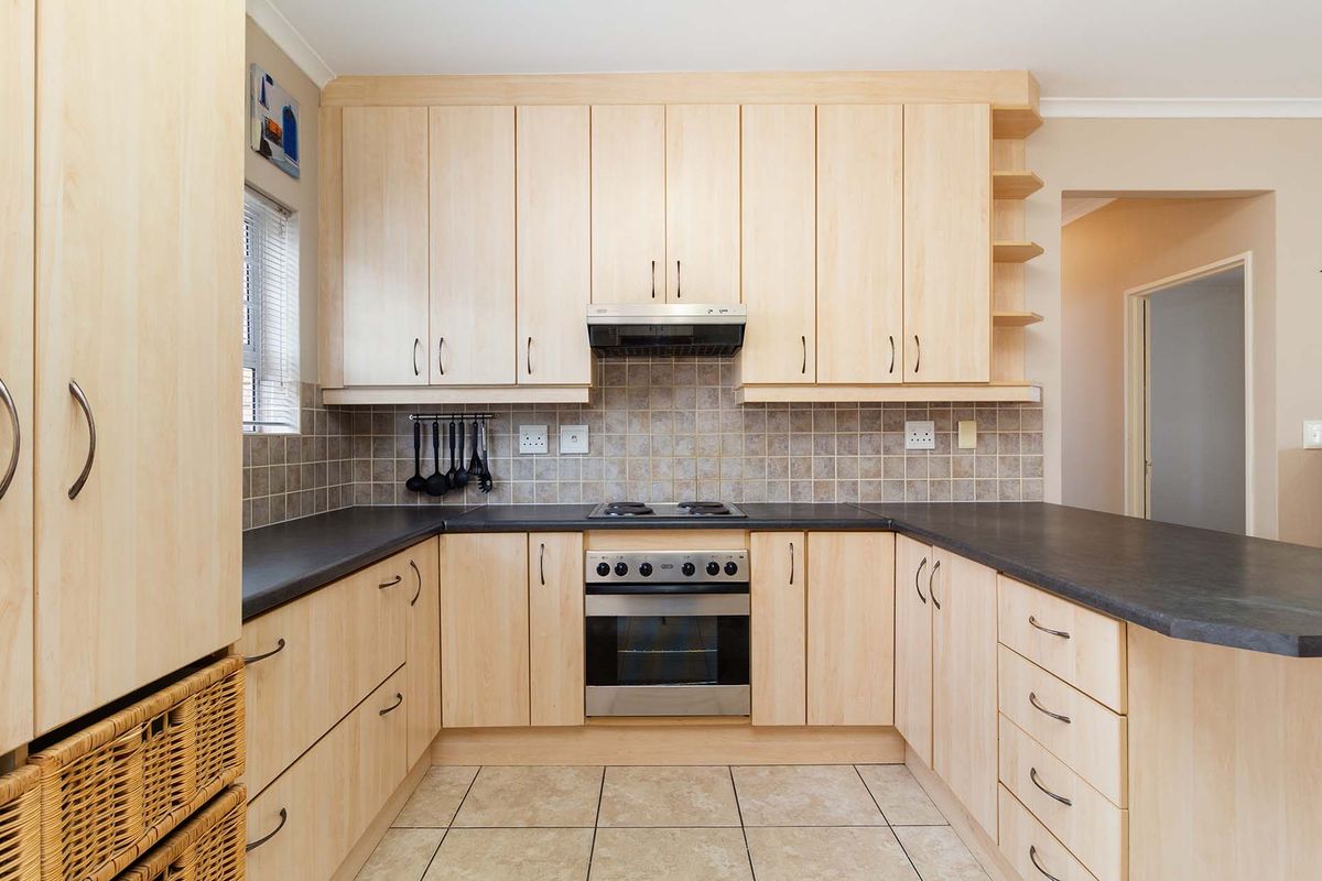 A functional kitchen featuring light wood-finish cabinetry, dark laminate countertops, a built-in oven with gas hob, and neutral tiled flooring and backsplash.