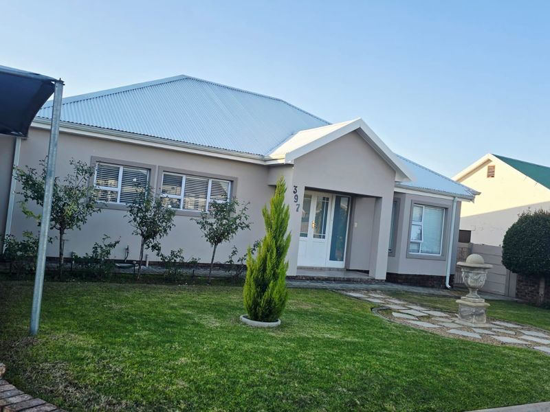 Front elevation of a well-maintained single-story house featuring a corrugated metal roof, paved driveway, carport, and landscaped front garden with a stone pathway.