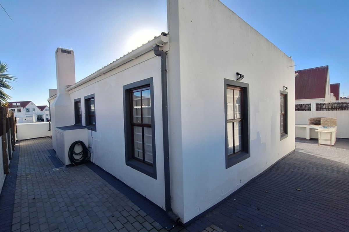 Rear exterior view of the property featuring a paved patio area, built-in braai, and clean white facade with dark window frames.