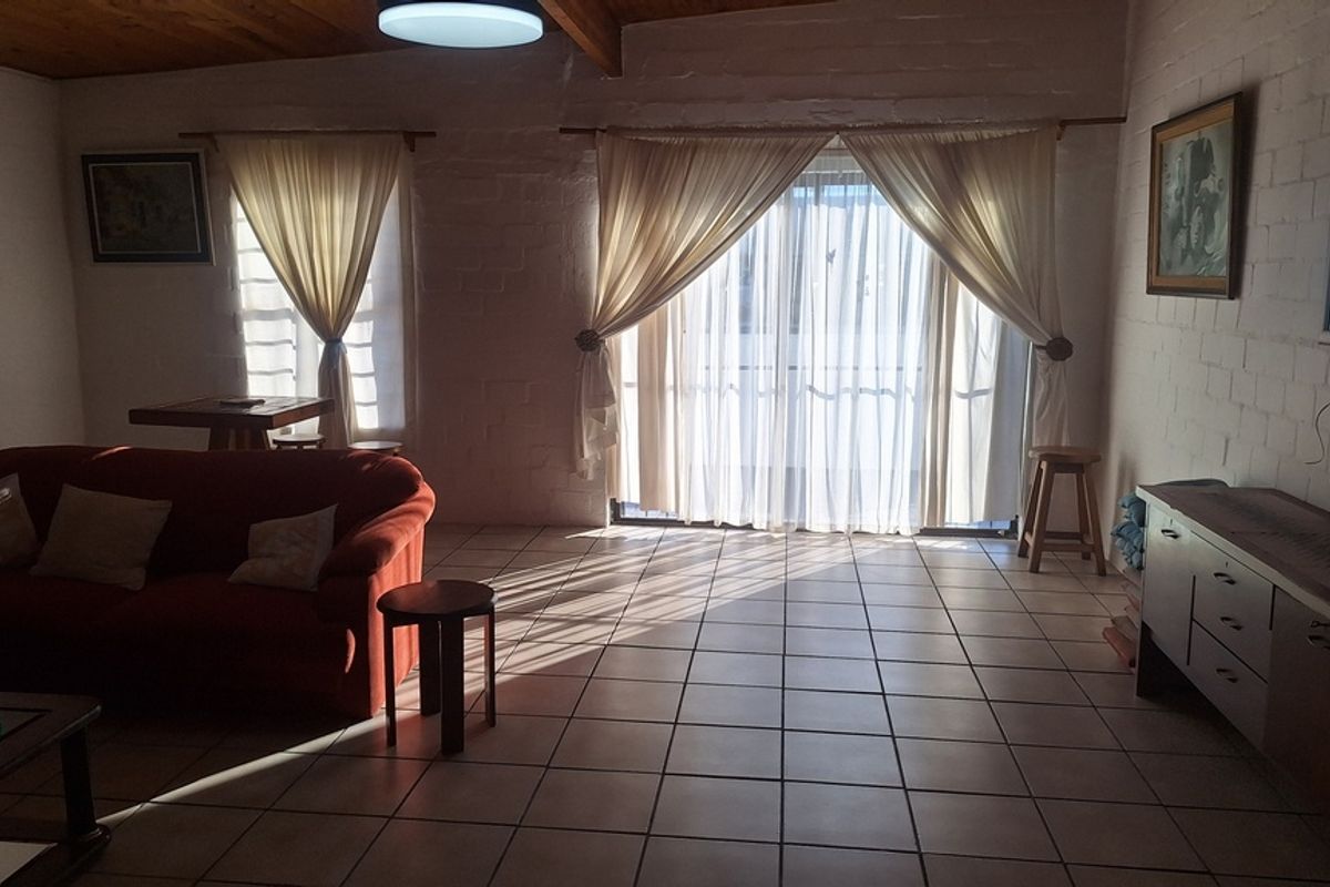 Spacious living room featuring exposed timber ceiling beams, ceramic floor tiles, and painted brick walls with direct access to an outdoor area via sliding doors.