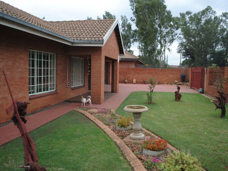 A view of the rear exterior featuring a brick house facade, paved pathways, a landscaped garden area, and secure perimeter walling.