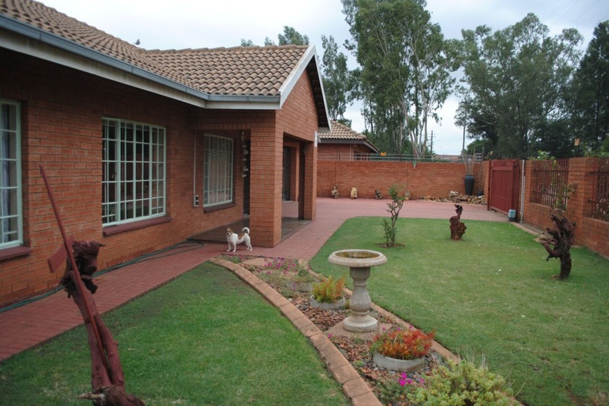A view of the rear exterior featuring a brick house facade, paved pathways, a landscaped garden area, and secure perimeter walling.