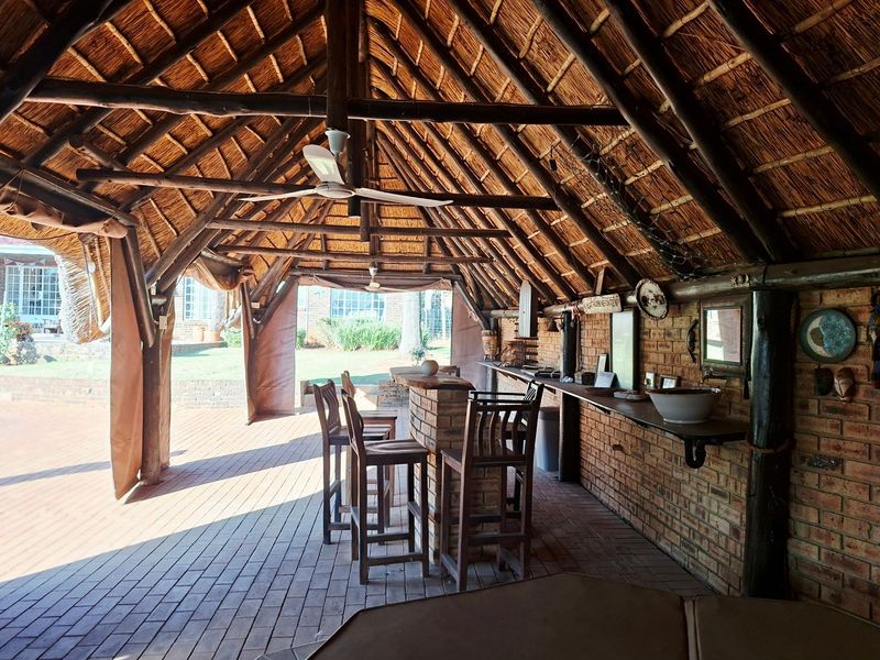 A spacious covered outdoor entertainment area featuring a thatched roof, exposed timber trusses, and a built-in brick bar counter overlooking the garden.