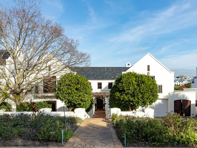 A well-maintained white rendered residence featuring a brick paved entrance path, slate roof, and integrated garage, set within a landscaped garden.