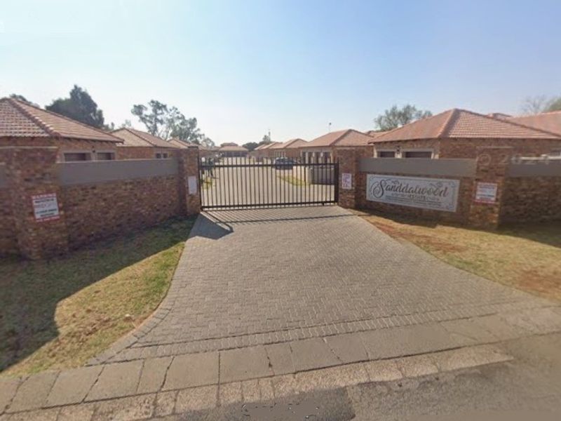 The entrance to Sanddalwood estate featuring a secure metal gate, brick perimeter walls, and a paved driveway leading into the townhouse complex.