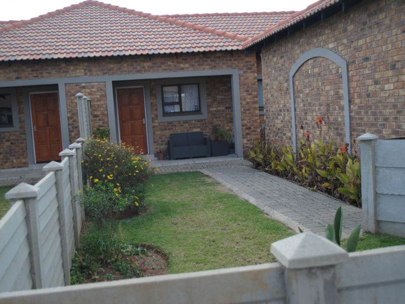 A well-maintained townhouse rear exterior featuring a face brick facade, tiled roof, paved walkway, and a private garden area with a covered patio.