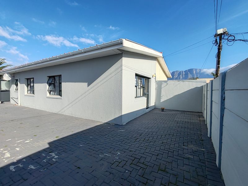 Rear exterior view of the property featuring a paved courtyard area, precast boundary walling, and aluminium window frames under a clear sky.