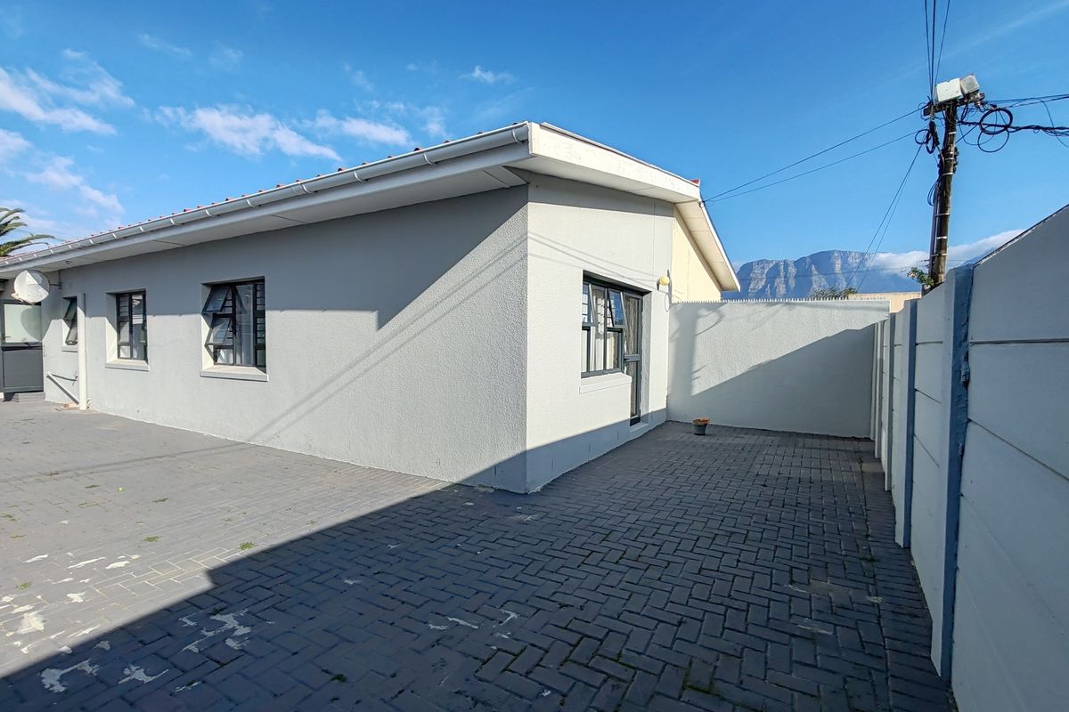 Rear exterior view of the property featuring a paved courtyard area, precast boundary walling, and aluminium window frames under a clear sky.