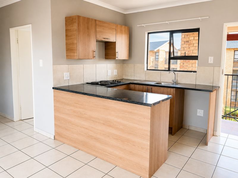 A clean, modern kitchen featuring light wood cabinetry, dark granite countertops, an integrated electric hob, and neutral tiled flooring with direct balcony access.