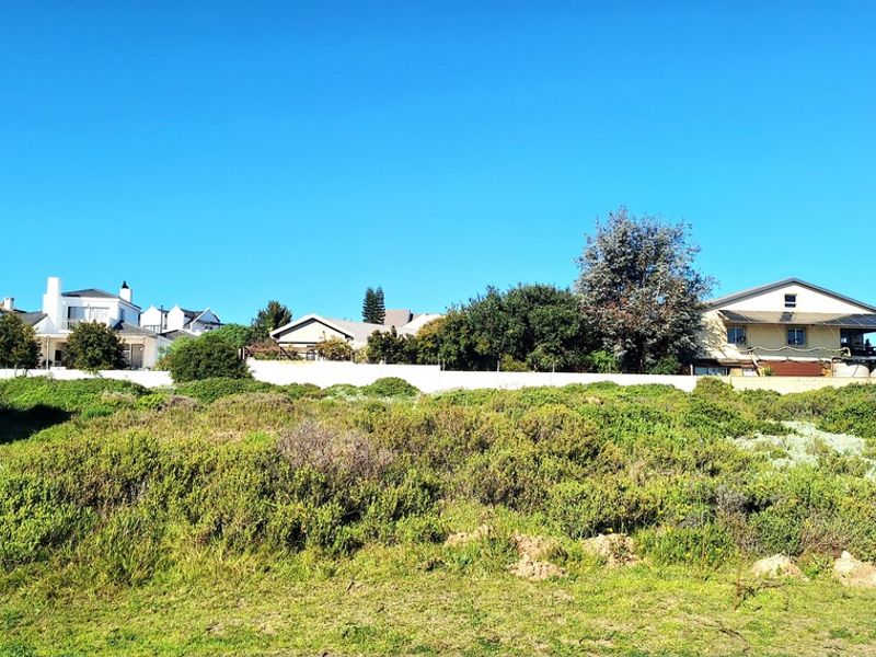 A view of a vacant residential plot featuring natural vegetation and a boundary wall with neighboring houses in the background.