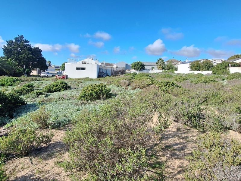 A vacant residential plot featuring sandy terrain and native coastal vegetation, situated in a developed neighborhood setting.