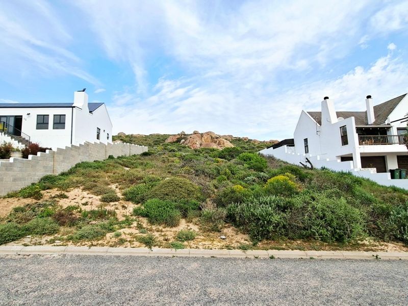 A vacant residential plot situated between two existing homes, featuring natural coastal vegetation and a concrete retaining wall.