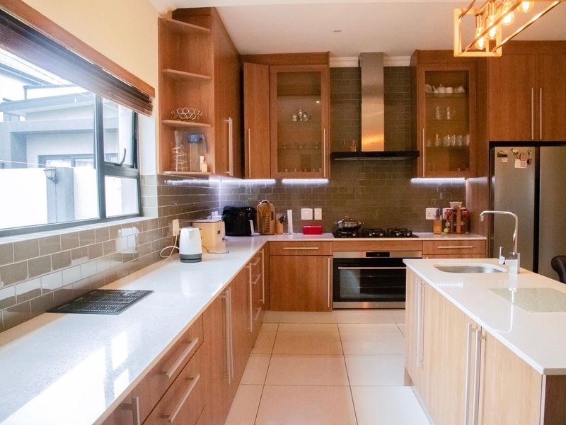 Modern kitchen featuring wood-finish cabinetry, stone countertops, a central island with sink, and integrated stainless steel appliances with stylish under-cabinet lighting.