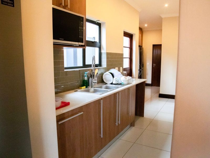 A functional kitchen featuring wood-finish cabinetry, a double sink with mixer tap, tiled backsplash, and built-in microwave, leading into a hallway.
