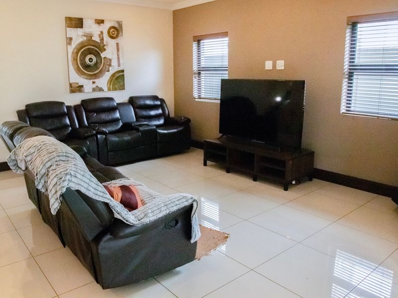 A spacious living room featuring polished floor tiles, neutral wall tones, and dual windows fitted with venetian blinds.