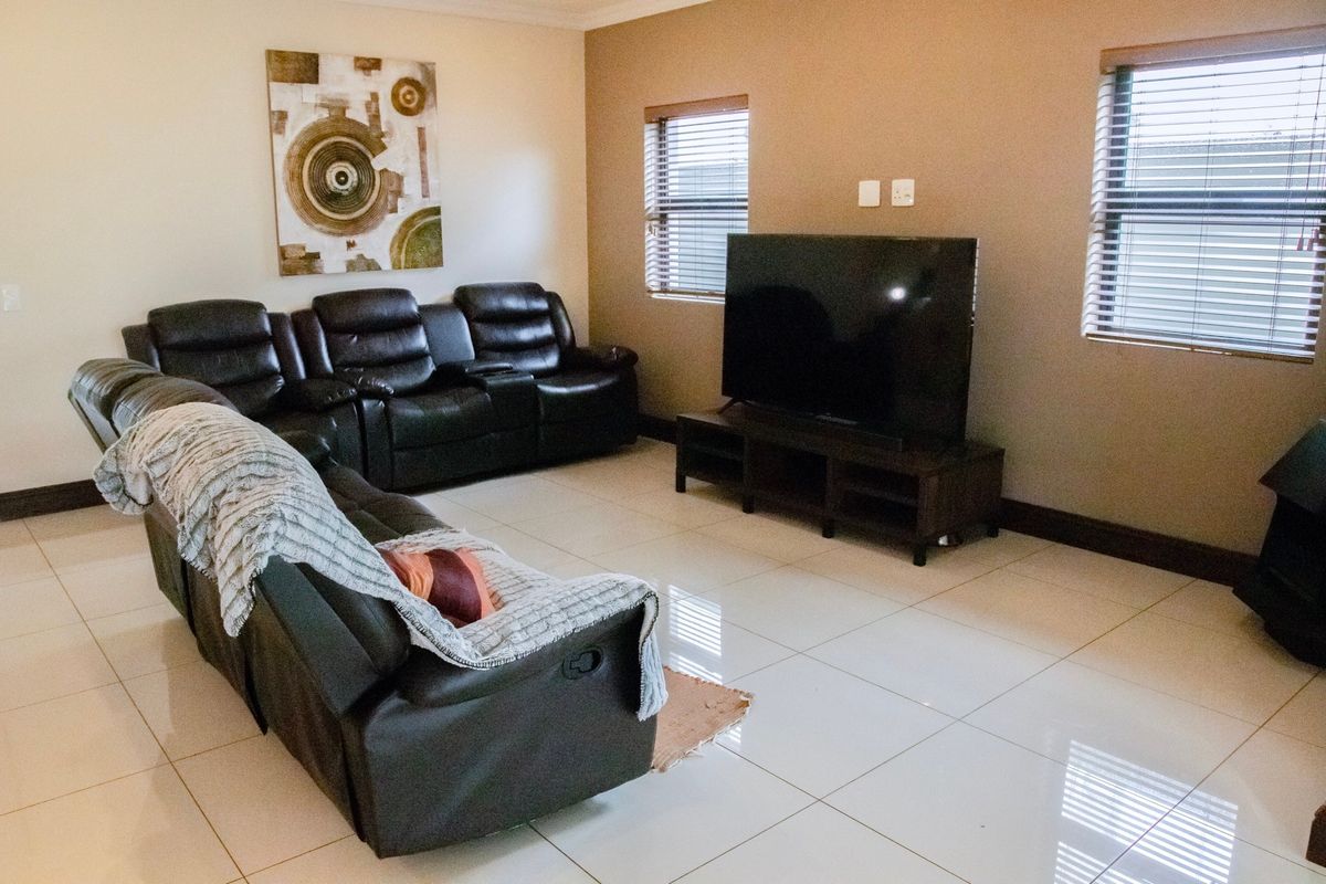 A spacious living room featuring polished floor tiles, neutral wall tones, and dual windows fitted with venetian blinds.