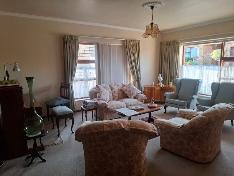 A traditional living room featuring carpeted flooring, a decorative ceiling rose with a pendant light, and large windows allowing natural light.