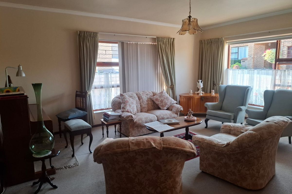 A traditional living room featuring carpeted flooring, a decorative ceiling rose with a pendant light, and large windows allowing natural light.