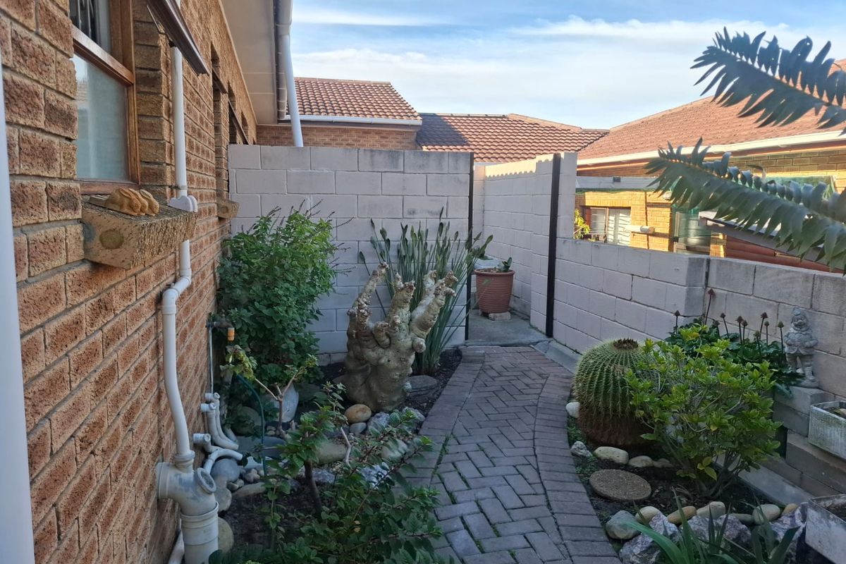 A side garden pathway featuring brick paving, established landscaping, and a cinder block boundary wall along the side of the townhouse.