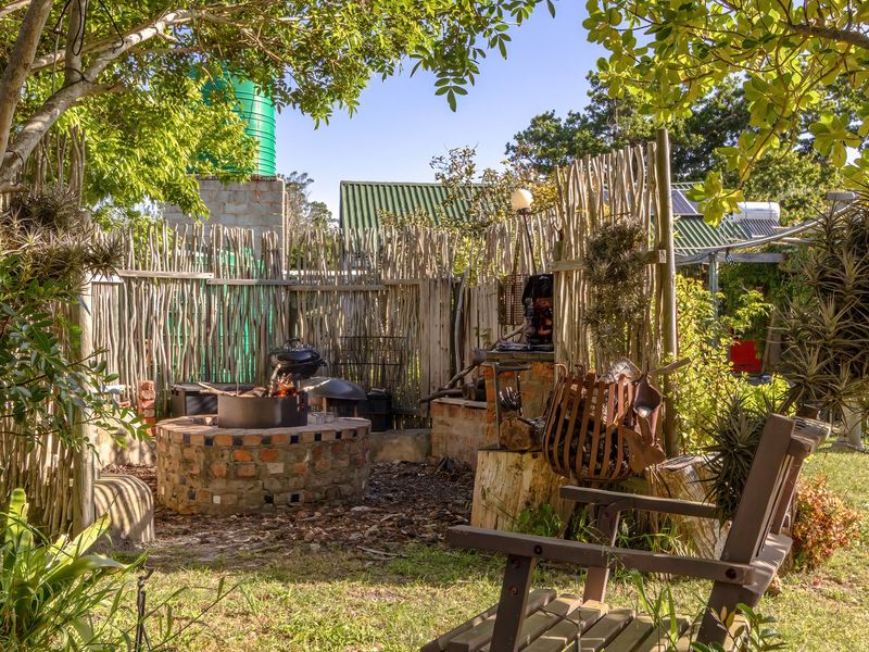 A rustic outdoor garden area featuring a brick fire pit, built-in braai infrastructure, and natural wooden pole fencing, set within a lush, established garden.